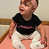 baby, infant, child, smile, headband, pink, black_shirt, metallica, blue_eyes, sitting, bed, pink_sheet, white_pants, baby_shoes, bracelet, cute, happy, indoors, portrait, person