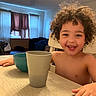 Elijah is registered to the contest to win money with this photo: child, smiling, curly_hair, portrait, indoor, home, table, cups, bowl, breakfast, sitting, happy, face, hands, wooden_table, window, curtains, chair, casual, family