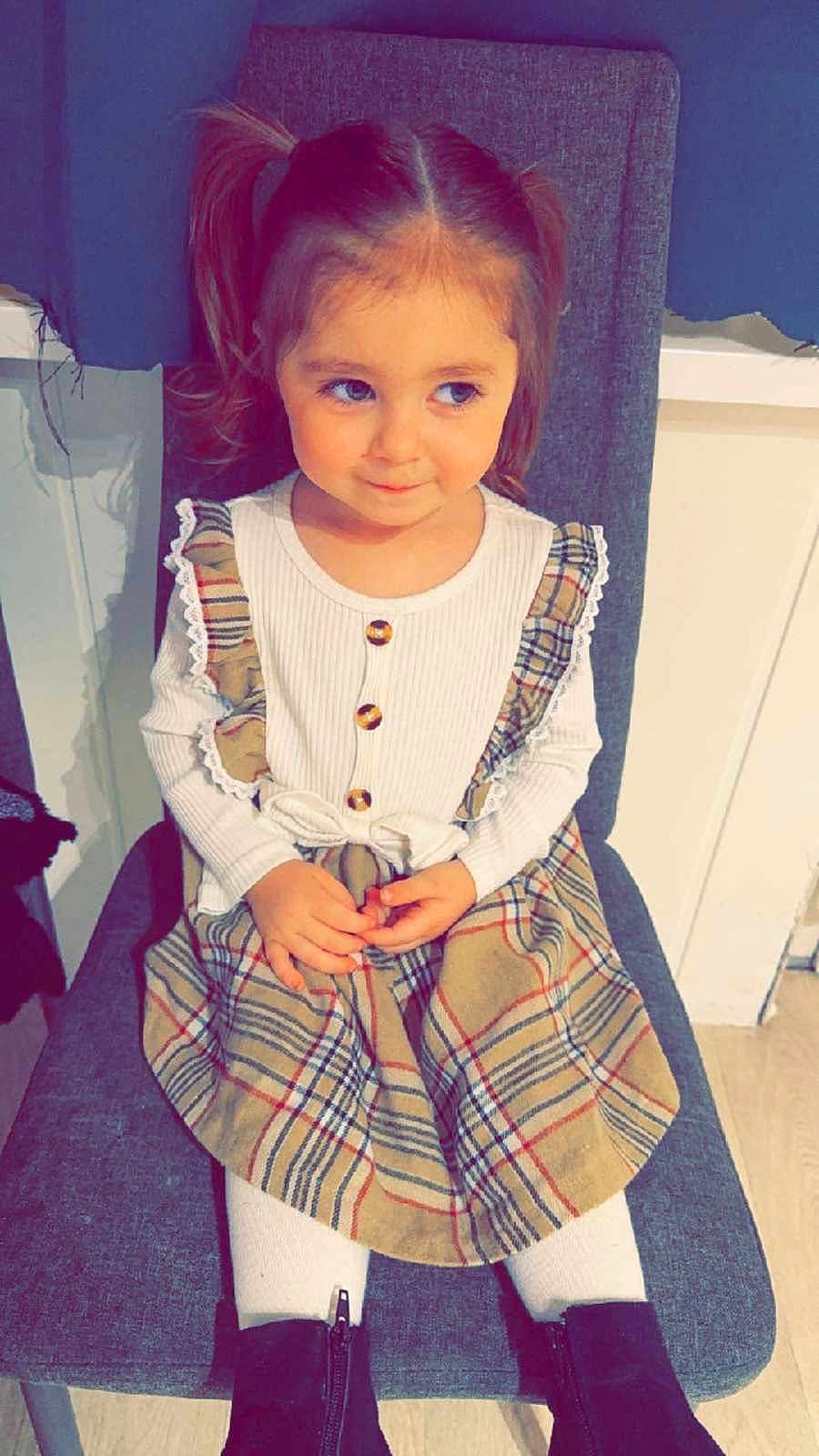 Stella participe au concours pour gagner de l'argent avec cette photo : child, girl, pigtails, plaid_dress, white_tights, black_boots, chair, indoor, smile, cute, sitting, long_sleeve_shirt, buttons, hands, floor, wall, furniture, portrait, young_child, happy