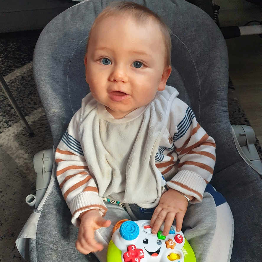 Shayron a rejoint le concours — aidez-le/la à gagner de superbes lots ! baby, bib, blue_eyes, carpet, child, curious, cute, face, game_controller, gray_chair, hand, home, indoor, infant, person, sitting, smiling, striped_clothing, toy, young