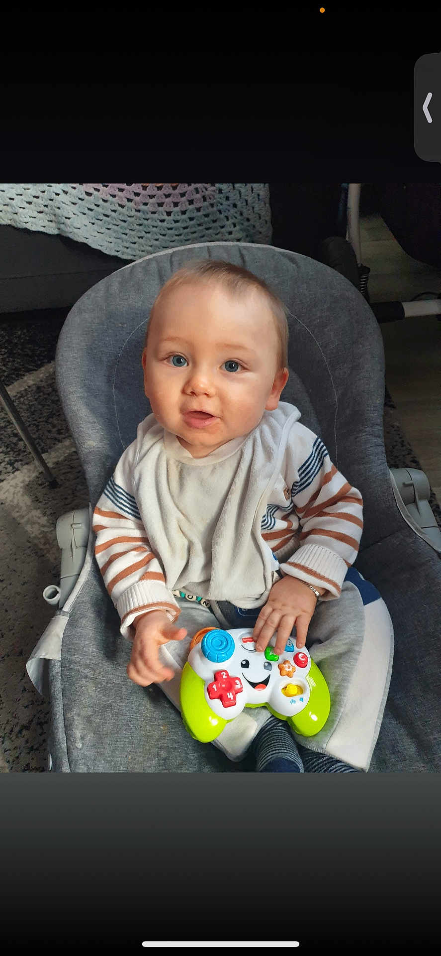 Shayron a rejoint le concours — aidez-le/la à gagner de superbes lots ! baby, child, infant, toy, game_controller, striped_clothing, bib, sitting, gray_chair, indoor, carpet, curious, blue_eyes, hand, face, smiling, young, cute, person, home