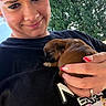 person, puppy, dog, sleeping, cuddling, portrait, selfie, outdoor, trees, nature, smiling, face, hands, black_shirt, brown_fur, small_dog, pet, adorable, manicure, daylight