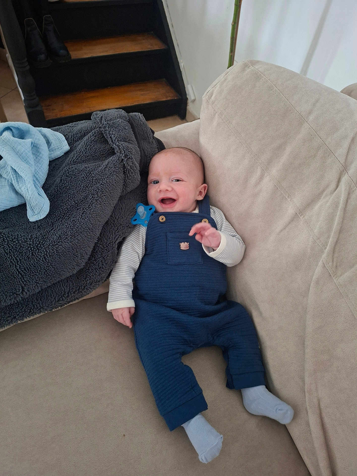Gaspard participe au concours pour gagner de l'argent avec cette photo : baby, infant, smile, couch, blanket, pacifier, overalls, socks, living_room, staircase, wood_stairs, plush_blanket, happy, sitting, portrait, clothing, person, furniture, cozy, cute