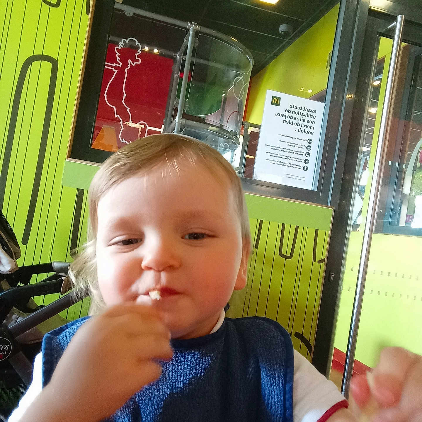 Yanael participe au concours pour gagner de l'argent avec cette photo : baby, bib, child, clothing, colorful, eating, face, fast_food, food, french_fries, glass, hand, high_chair, indoor, person, play_area, restaurant, smile, toddler, window