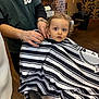 Adem Sofiane participe au concours pour gagner de l'argent avec cette photo : barber, barber_cape, bracelet, brick_wall, chair, child, curly_hair, floor, furniture, haircut, haircut_session, hands, indoor, lamp, person, plant, salon, striped_cape, toddler, wide_eyes