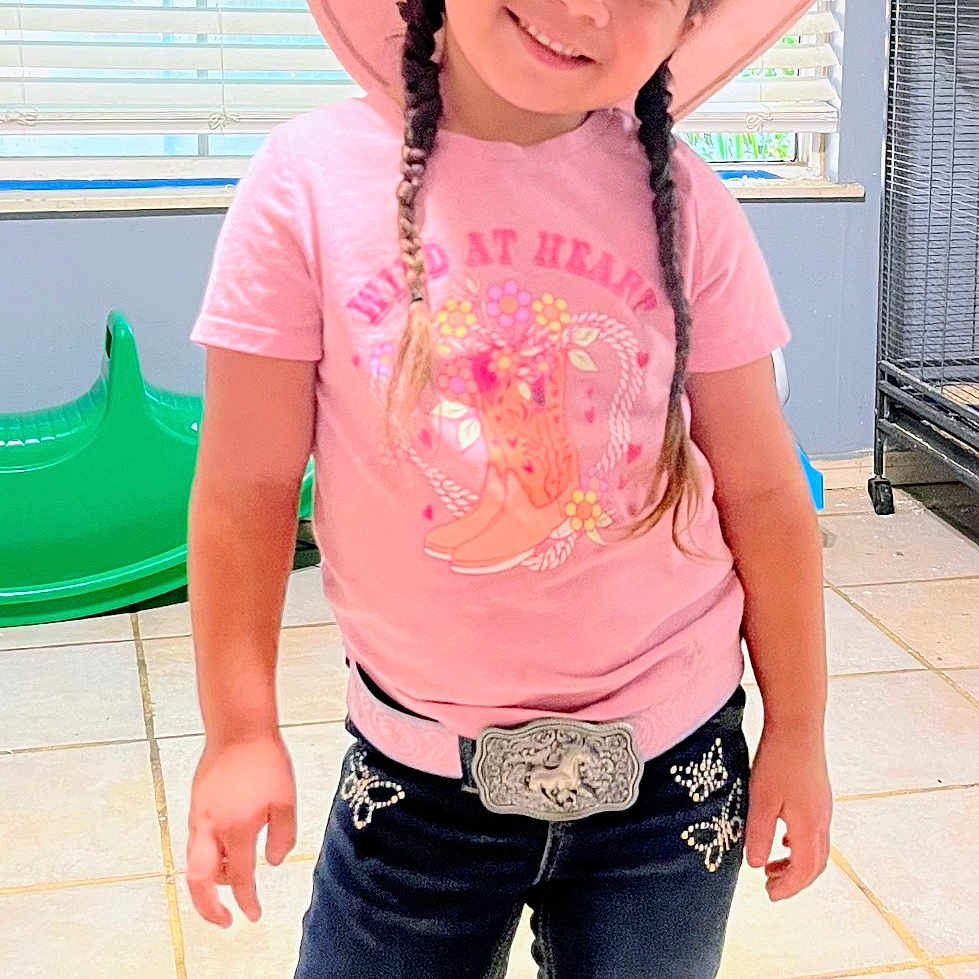 Zaela is registered to the contest to win money with this photo: accessory, belt_buckle, blinds, braids, child, cowboy_boots, cowgirl, girl, happy, home_interior, indoor, jeans, pink_hat, playful, playroom, portrait, smile, standing, t_shirt, tile_floor