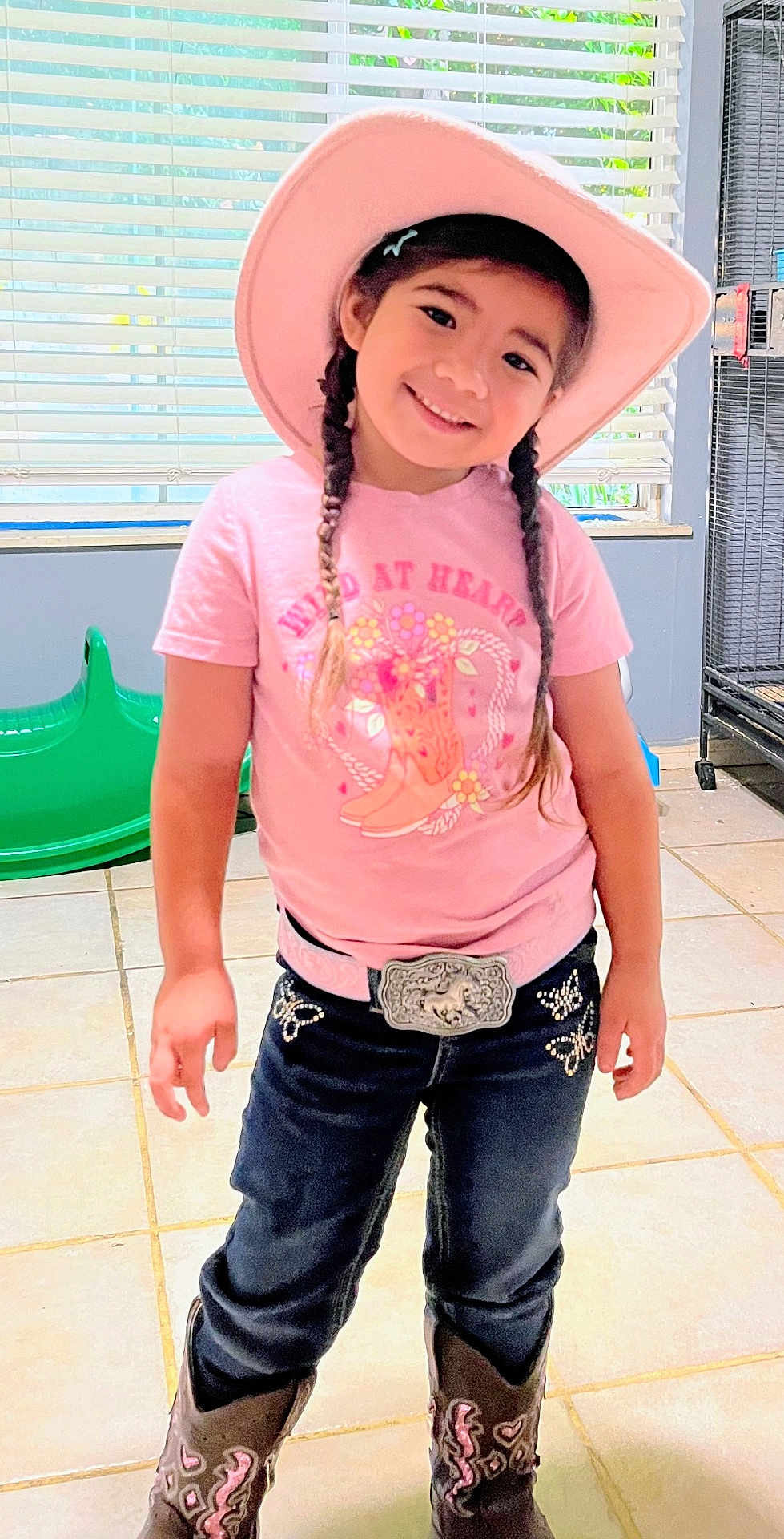 Zaela is registered to the contest to win money with this photo: child, girl, cowgirl, pink_hat, braids, smile, belt_buckle, cowboy_boots, jeans, t_shirt, indoor, blinds, tile_floor, playroom, happy, standing, portrait, accessory, playful, home_interior