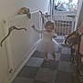 baby_gate, barefoot, checkered_floor, child, curtain, floor_tiles, hallway, hanger, home, indoor, joyful, person, radiator, smiling, stroller, toddler, toy, walking, white_dress, window