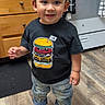 Sebastian is registered to the contest to win money with this photo: child, toddler, boy, smiling, portrait, standing, kitchen, oven, cabinets, wooden_floor, black_shirt, burger_shirt, jeans, sneakers, short_hair, indoors, home, happy, full_body, photograph