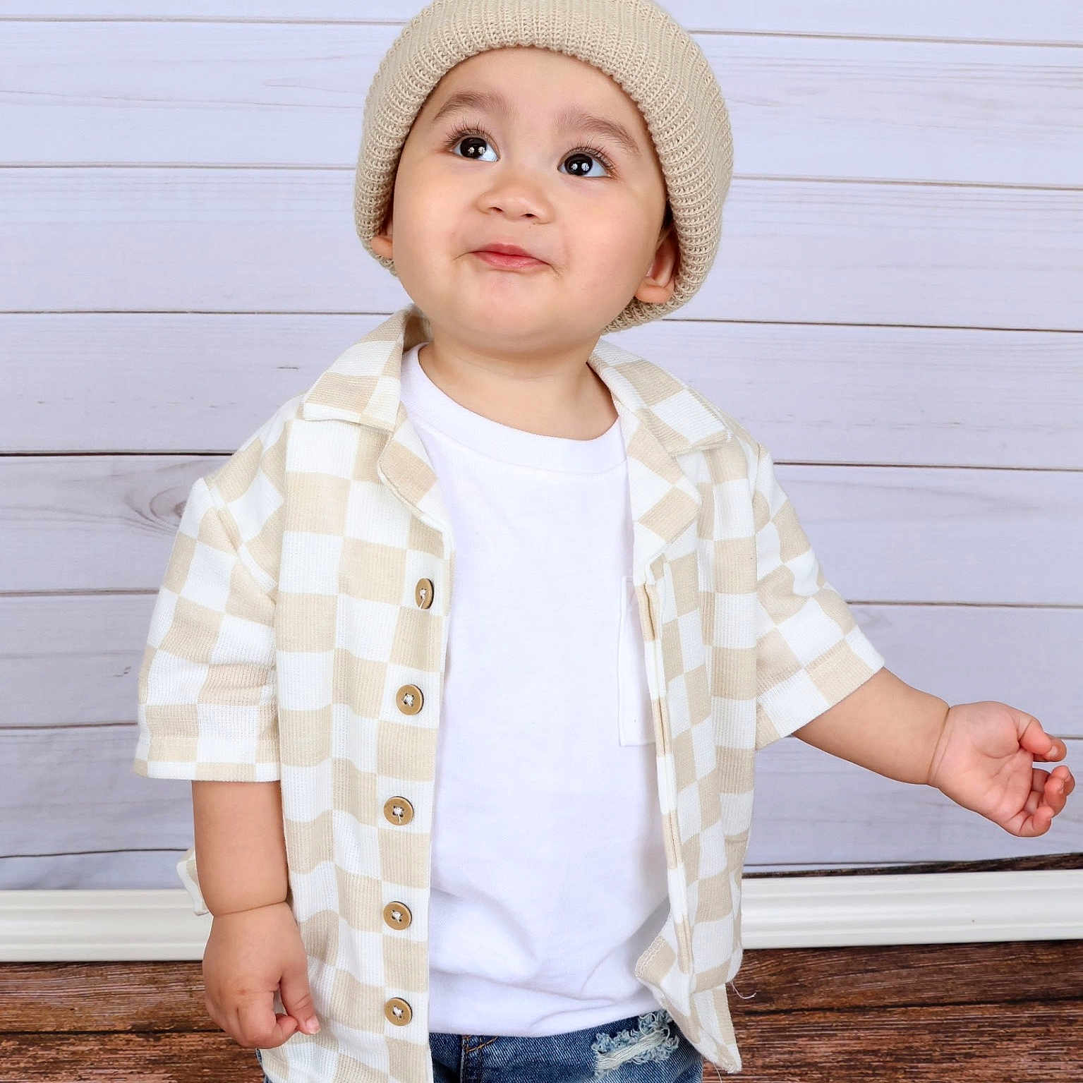 Sebastian is registered to the contest to win money with this photo: baby, child, toddler, portrait, standing, fashion, beanie, hat, shirt, checkered_pattern, t_shirt, jeans, distressed_denim, outdoor_style, wooden_background, studio_portrait, smiling, face, person, photography
