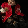 Sebastian is registered to the contest to win money with this photo: child, toddler, baby, portrait, smiling, sitting, beanie, red_jacket, varsity_jacket, camouflage_pants, black_clothing, fashion, indoor, dark_background, chair, face, eyes, happy, casualwear, photography