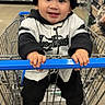 child, toddler, baby, smiling, happy, portrait, shopping_cart, supermarket, grocery_store, indoor, beanie, baseball_jersey, black_clothing, white_clothing, sneakers, cart_handle, sitting, shopping, shelves, retail