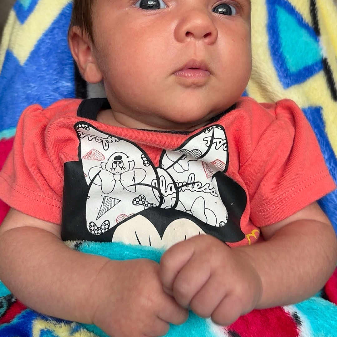 Oak is registered to the contest to win money with this photo: baby, blanket, bow, clothing, colorful, cozy, cute, expression, face, hands, indoor, infant, orange_shirt, person, portrait, seated, soft_texture, wide_eyes, wrapped, young_child