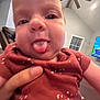 Oak is registered to the contest to win money with this photo: baby, tongue, close_up, face, hand, maroon_clothing, patterned_clothing, ceiling_fan, window, television, cartoon, indoor, person, child, playful, smiling, skin, head, expression, home