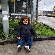 Swan participe au concours pour gagner de l'argent avec cette photo : toddler, child, smiling, curly_hair, black_jacket, jeans, sneakers, bread, outdoor, urban, sidewalk, plants, flowers, building, glass_window, lamp_post, sky, greenery, happy, sitting