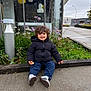 toddler, child, smiling, curly_hair, black_jacket, jeans, sneakers, bread, outdoor, urban, sidewalk, plants, flowers, building, glass_window, lamp_post, sky, greenery, happy, sitting