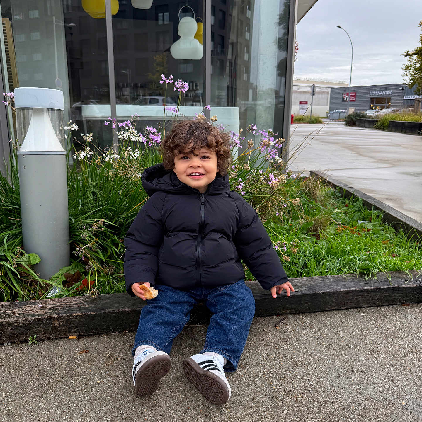 Swan participe au concours pour gagner de l'argent avec cette photo : toddler, child, smiling, curly_hair, black_jacket, jeans, sneakers, bread, outdoor, urban, sidewalk, plants, flowers, building, glass_window, lamp_post, sky, greenery, happy, sitting