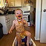 Kira is registered to the contest to win money with this photo: child, toddler, baby, messy, food, baby_food, diaper, booster_seat, kitchen, stove, cabinets, wooden_floor, bowl, cup, spoon, laughing, smiling, amber_necklace, face, hands