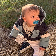 Alessio a rejoint le concours — aidez-le/la à gagner de superbes lots ! baby, casual_clothing, child, cold_weather, curious, daylight, footwear, grass, gravel, jacket, nature, outdoor, pacifier, portrait, puffy_jacket, shadow, sitting, sunlight, toddler, young_child