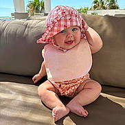 Freya is registered to the contest to win money with this photo: baby, child, hat, bib, smile, outdoor, sofa, cushion, sunlight, happy, blue_eyes, legs, feet, skin, clothing, summer, vacation, palm_trees, relaxation, portrait