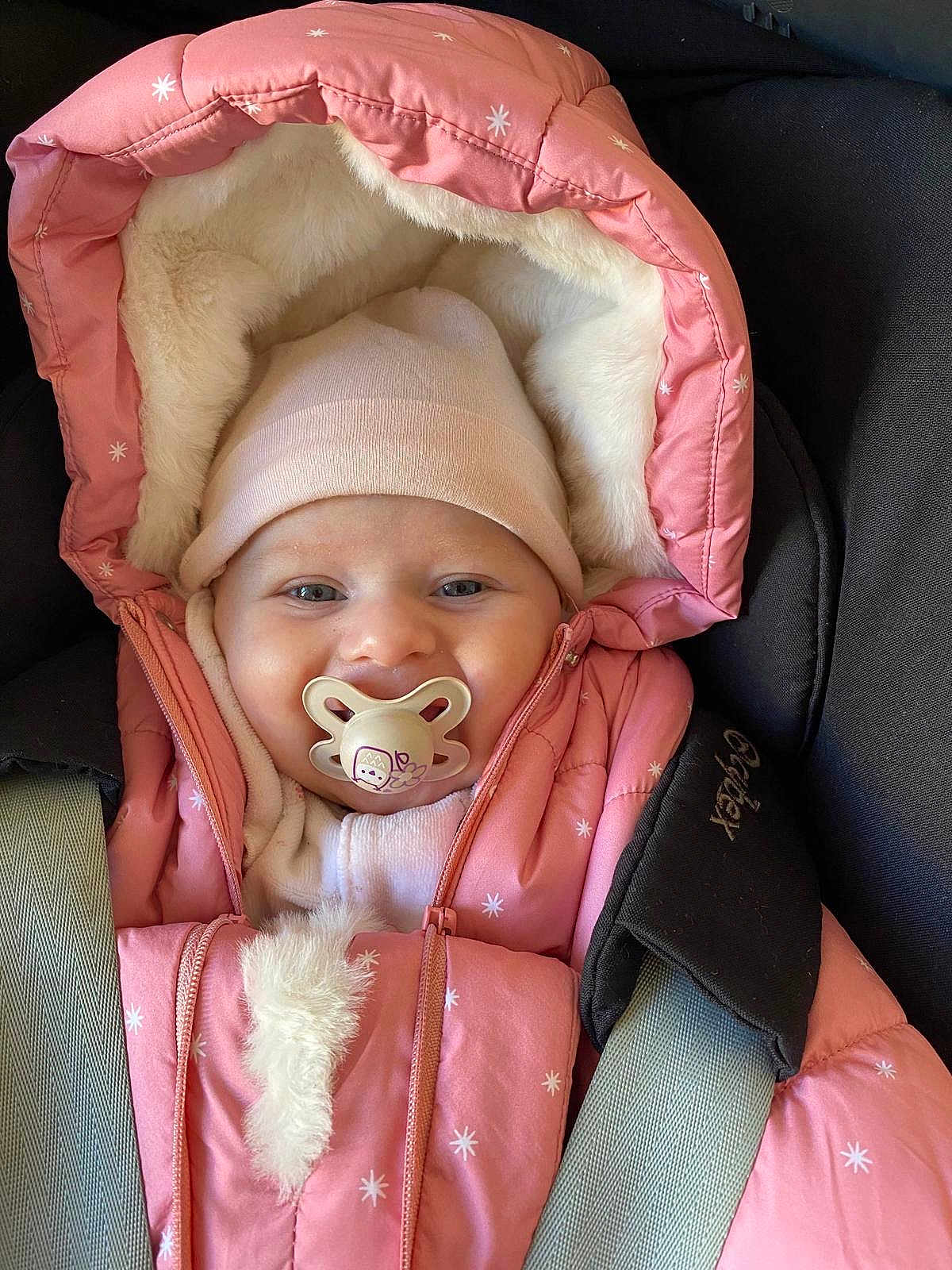 Manon a rejoint le concours — aidez-le/la à gagner de superbes lots ! baby, pacifier, pink_jacket, hat, car_seat, child, winter_clothing, fur_lining, seatbelt, cute, infant, portrait, warm, smiling_eyes, cozy, indoor, closeup, face, small_child, adorable