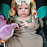 baby, child, headband, bow, car_seat, plush_toy, purple_toy, green_clothing, floral_pillow, infant, cute, portrait, indoors, serious_expression, child_in_car_seat, soft_toy, sitting, toddler, baby_clothes, baby_face