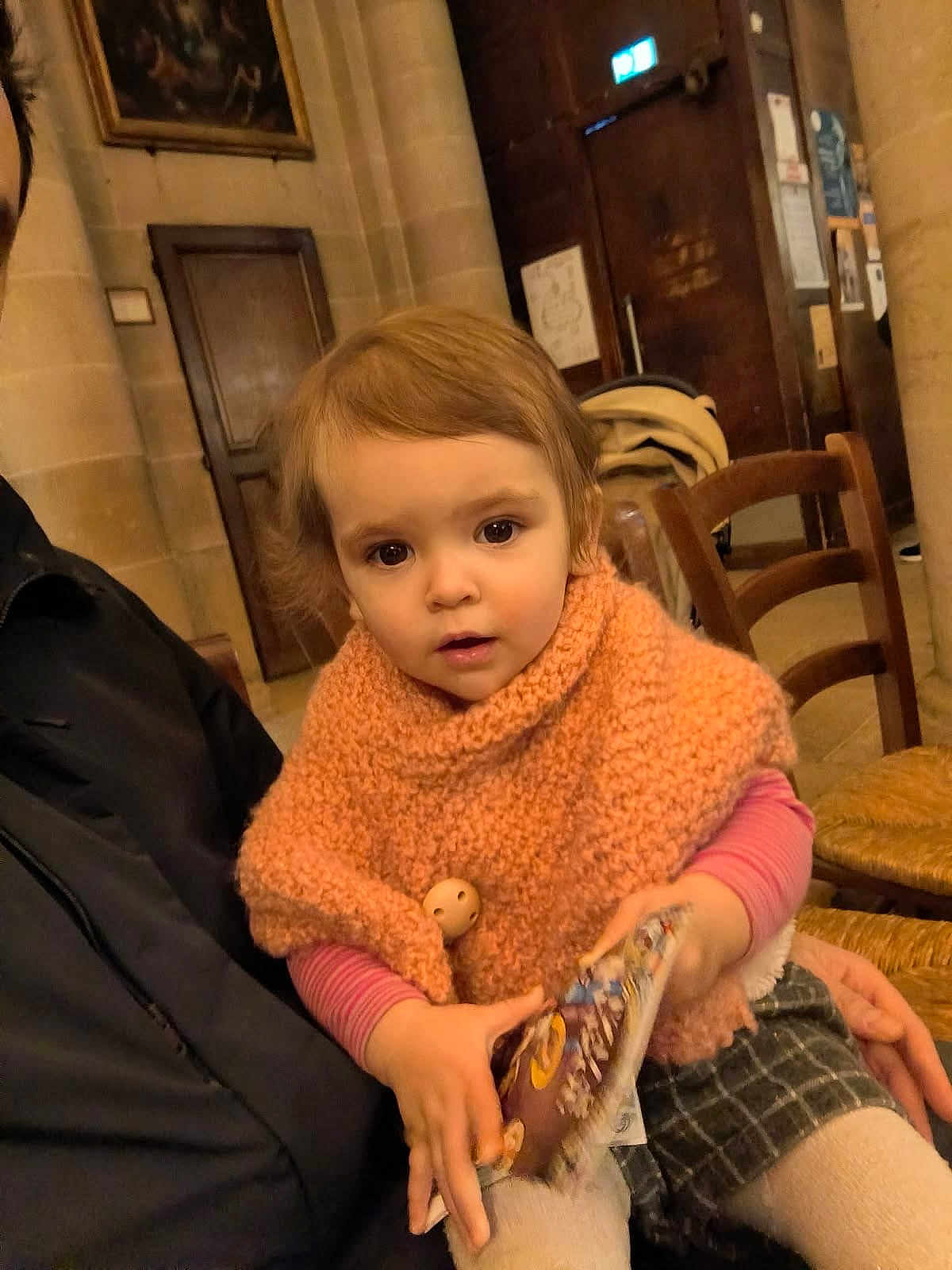 Camille participe au concours pour gagner de l'argent avec cette photo : toddler, child, person, indoor, knit_poncho, orange_clothing, book, seated, adult, hand, wooden_chair, stone_wall, warm_lighting, curly_hair, plaid_skirt, white_tights, portrait, curious, holding_object, casual_clothing