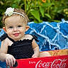baby, child, face, headband, flower, blue_eyes, black_clothing, crate, red, coca_cola, outdoor, greenery, blanket, smile, portrait, cute, summer, nature, infant, happy