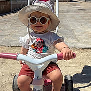 Alexane participe au concours pour gagner de l'argent avec cette photo : building, child, footwear, fun, handlebars, hat, outdoor, pavement, play, shadow, shorts, smiling, summer, sunglasses, sunny, toddler, toy, tree, tricycle, tshirt