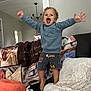 toddler, child, jumping, blue_sweatshirt, shorts, curly_hair, smiling, living_room, sofa, blanket, pillow, indoor, happy, excited, home, person, couch, furniture, playful, cheerful