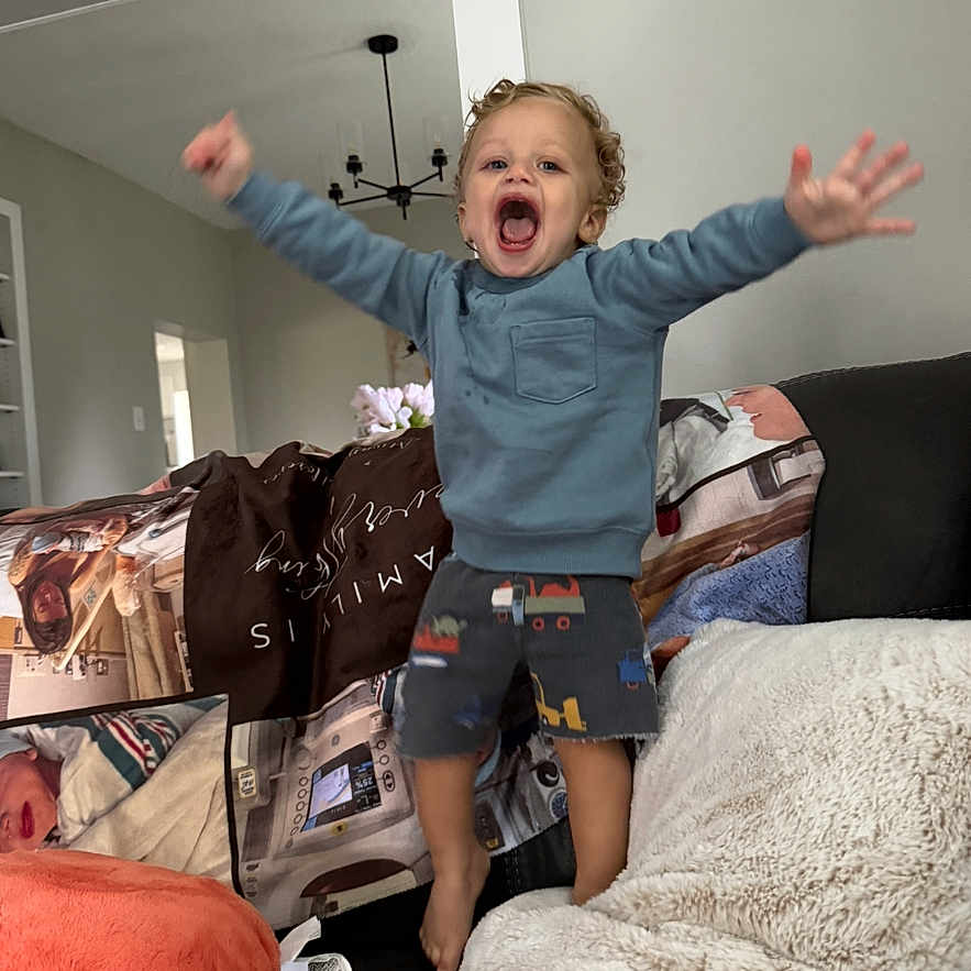 Malachi is registered to the contest to win money with this photo: blanket, blue_sweatshirt, cheerful, child, couch, curly_hair, excited, furniture, happy, home, indoor, jumping, living_room, person, pillow, playful, shorts, smiling, sofa, toddler