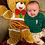 baby, teddy_bear, plush_toy, green_sweater, couch, blanket, holiday, festive, smile, child, indoor, cute, cozy, red_bow_tie, pants, patterned_clothing, furniture, happy, portrait, soft_toy