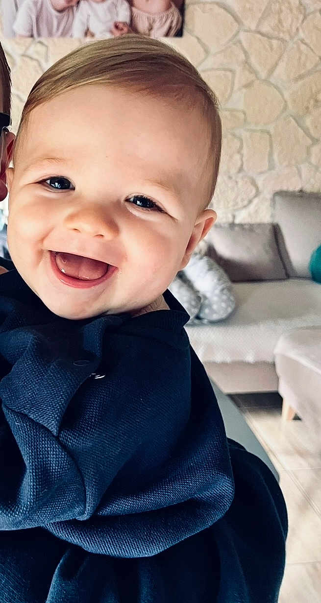 Julián participe au concours pour gagner de l'argent avec cette photo : baby, smiling, face, child, indoors, living_room, sofa, stone_wall, cozy, happiness, portrait, closeup, cute, apparel, person, home, furniture, cushion, sitting, joy
