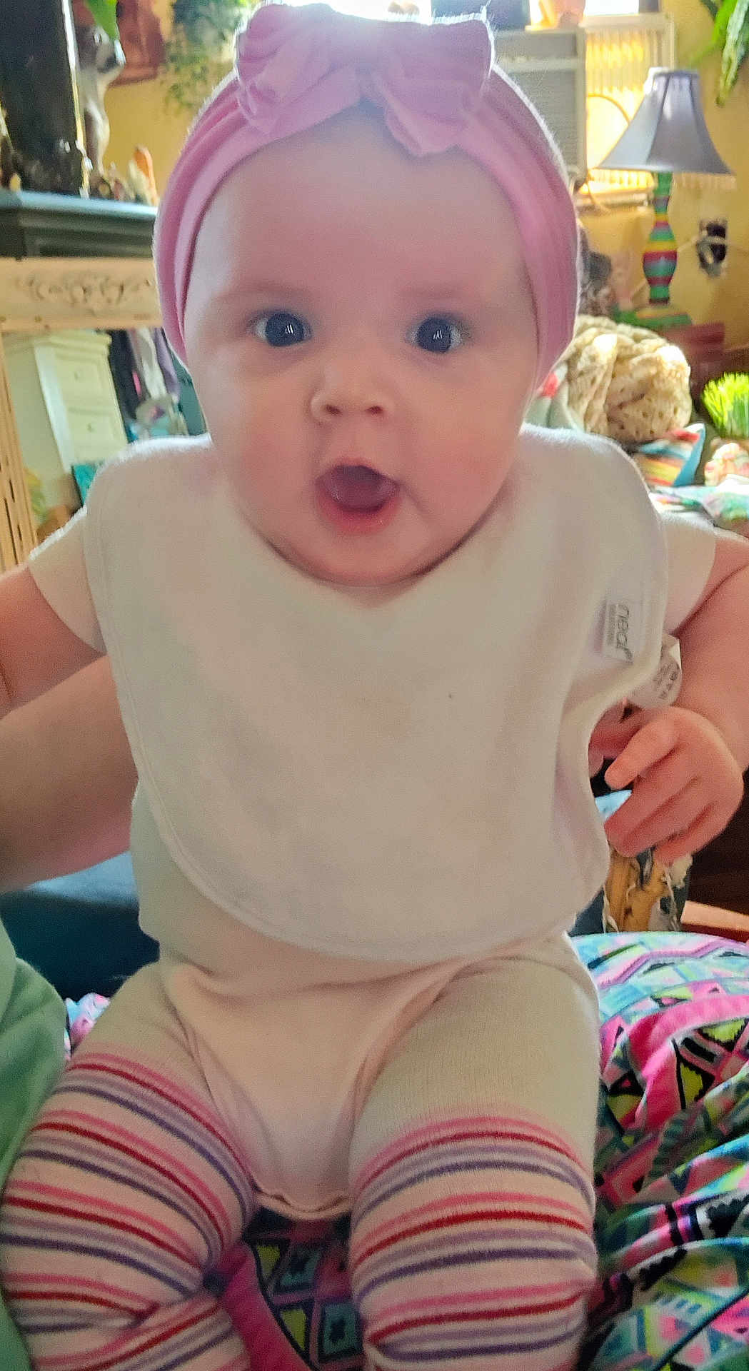 Lani is registered to the contest to win money with this photo: baby, infant, pink_headband, bib, striped_leggings, wide_eyes, surprised_expression, chubby_cheeks, cute, closeup, sitting, indoor, living_room, lamp, blanket, toy, furniture, portrait, colorful, person
