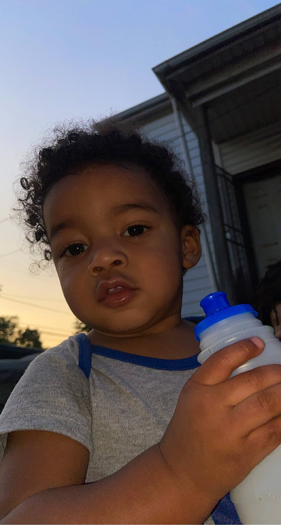 Kai is registered to the contest to win money with this photo: bottle, child, drinking_water, drinkware, electric_blue, facial_expression, fluid, gesture, hand, happy, mouth, nose, person, plastic_bottle, skin, sky, thumb, toddler, tree, water