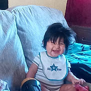 Tre'Mani joined the competition — help win amazing prizes! toddler, child, baby, smiling, sitting, couch, blanket, pillow, living_room, indoor, portrait, dark_hair, casual_clothing, bib, toy, red_toy, comfortable, happy, home, photograph