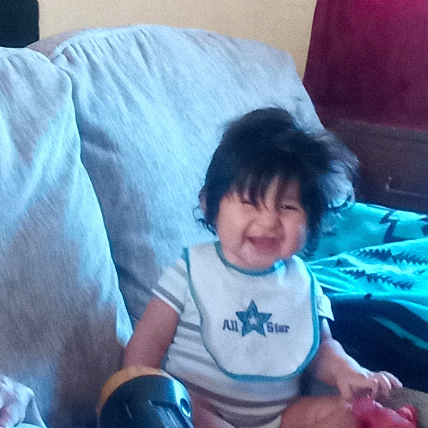 Tre'Mani joined the competition — help win amazing prizes! toddler, child, baby, smiling, sitting, couch, blanket, pillow, living_room, indoor, portrait, dark_hair, casual_clothing, bib, toy, red_toy, comfortable, happy, home, photograph