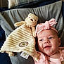 baby, smile, headband, pink_clothing, teddy_bear, stuffed_animal, blanket, cushion, fabric, indoor, child, soft_toy, happy, cute, infant, portrait, face, cozy, lying_down, toy