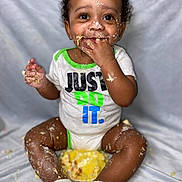 Kashmir is registered to the contest to win money with this photo: baby, child, toddler, sitting, portrait, smiling, happy, curly_hair, brown_skin, cake, food, messy, eating, hands, fingers, onesie, shirt, socks, indoor, fabric_backdrop