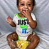 baby, child, toddler, sitting, portrait, smiling, happy, curly_hair, brown_skin, cake, food, messy, eating, hands, fingers, onesie, shirt, socks, indoor, fabric_backdrop