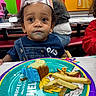child, toddler, birthday, party, crown, cake, frosting, fries, food, plate, sitting, portrait, curly_hair, smiling, indoor, restaurant, celebration, colorful, table, overalls
