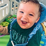 Walker is registered to the contest to win money with this photo: baby, child, smiling, green_shirt, blue_harness, playpen, toys, indoor, colorful, happy, cute, face, infant, person, young_child, play_area, expression, bright, fun, cheerful