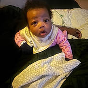 Regina is registered to the contest to win money with this photo: baby, infant, child, face, pink_clothing, bib, towel, blanket, quilt, hand, curly_hair, indoor, bed, candid, expression, wide_eyes, open_mouth, soft_texture, cozy, person