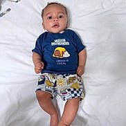 Sabira is registered to the contest to win money with this photo: baby, child, infant, blue_shirt, printed_shirt, shorts, lying_down, white_sheet, curly_hair, skin, face, hands, feet, indoors, colorful_clothing, expression, portrait, casual, cute, person