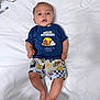 baby, child, infant, blue_shirt, printed_shirt, shorts, lying_down, white_sheet, curly_hair, skin, face, hands, feet, indoors, colorful_clothing, expression, portrait, casual, cute, person