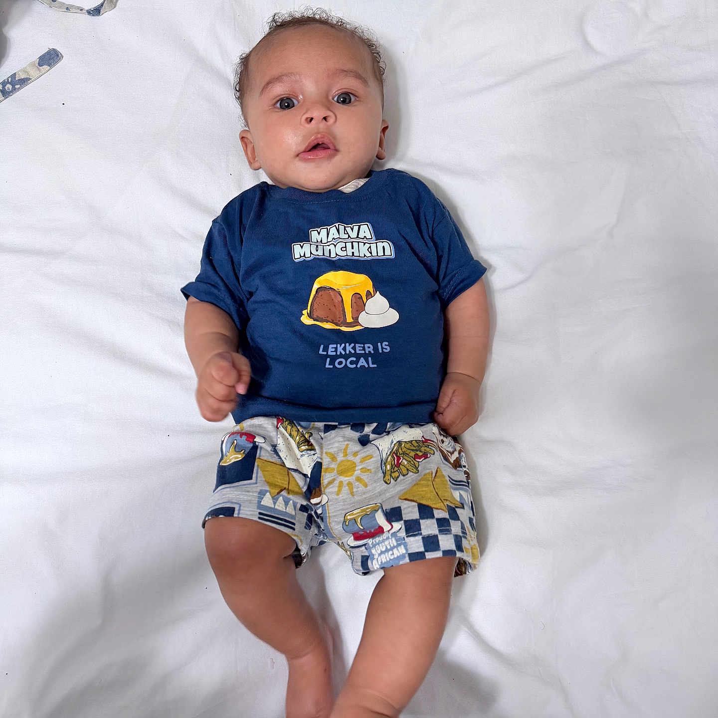 Sabira is registered to the contest to win money with this photo: baby, blue_shirt, casual, child, colorful_clothing, curly_hair, cute, expression, face, feet, hands, indoors, infant, lying_down, person, portrait, printed_shirt, shorts, skin, white_sheet