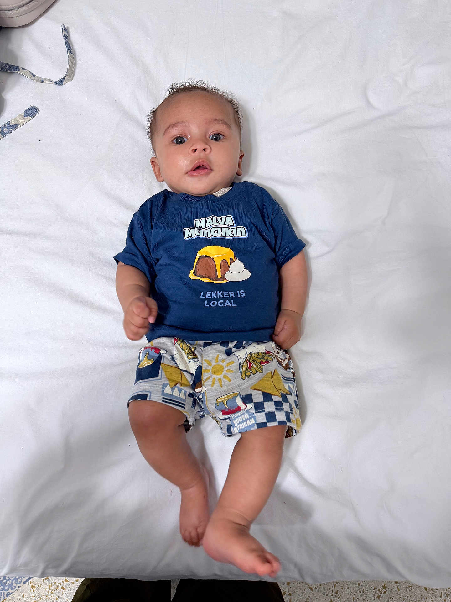 Sabira is registered to the contest to win money with this photo: baby, child, infant, blue_shirt, printed_shirt, shorts, lying_down, white_sheet, curly_hair, skin, face, hands, feet, indoors, colorful_clothing, expression, portrait, casual, cute, person