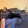 man, black_cat, couch, petting, indoor, relaxed, furniture, water_bottle, table, quilted_blanket, wooden_floor, shorts, tshirt, smiling, tail_up, domestic_cat, home, person, leisure, animal