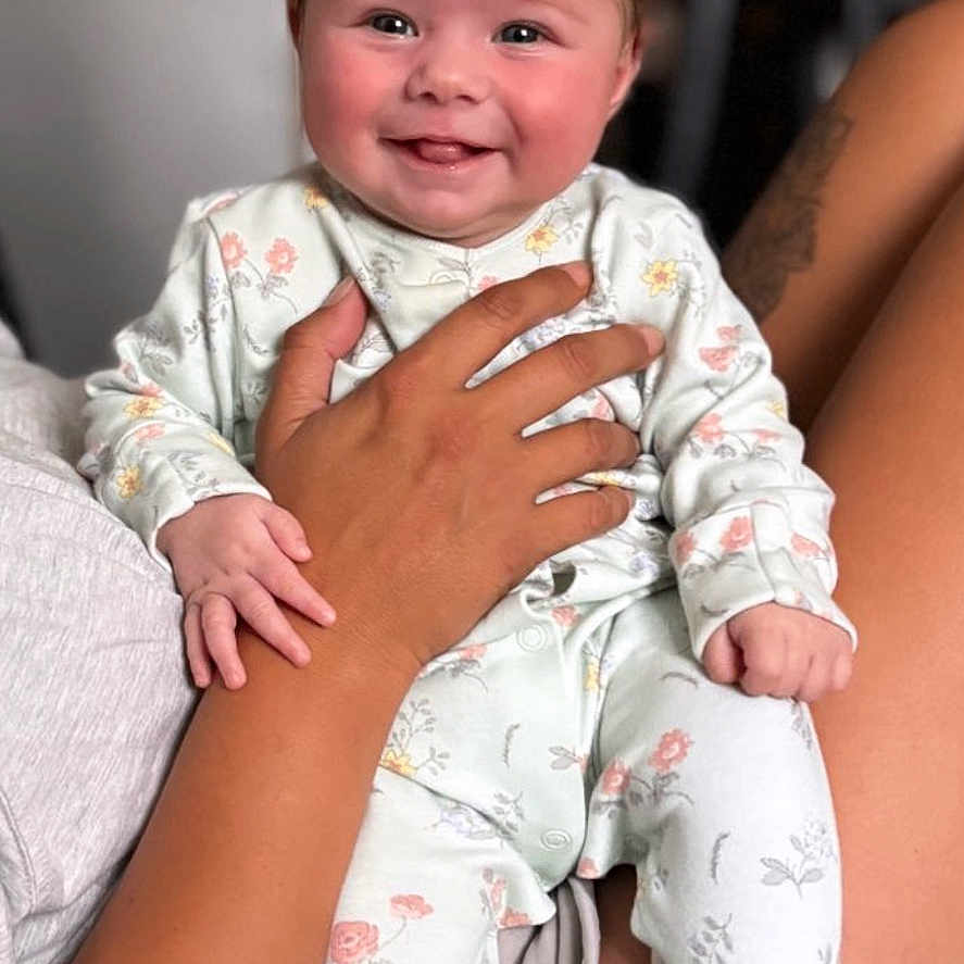Margot is registered to the contest to win money with this photo: baby, child, comfort, couch, cute, face, family, floral_pajamas, hand, happy, holding, indoor, infant, legs, person, portrait, sitting, skin, smiling, tattoo