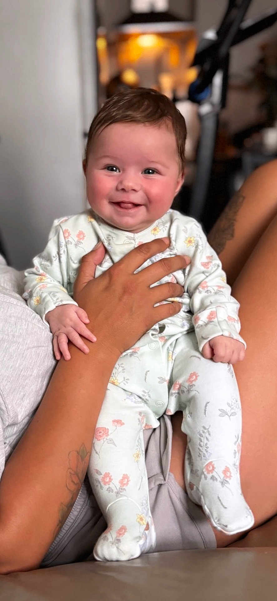 Margot is registered to the contest to win money with this photo: baby, child, smiling, floral_pajamas, tattoo, hand, indoor, person, couch, holding, happy, infant, face, legs, sitting, skin, comfort, portrait, cute, family
